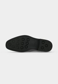 Semelle d'une chaussure habillée noire avec des crampons en caoutchouc ronds disposés en motif quadrillé et le nom de la marque ALDO visible près de la voûte plantaire.