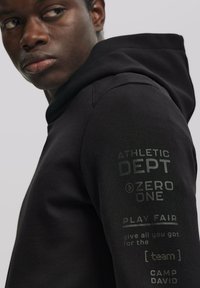 Fekete kapucnis pulóver, puha, sima anyagból készült. Az ujjon nyomtatott szöveg található, beleértve az "ATHLETIC DEPT" és a "PLAY FAIR" feliratokat.