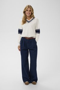 Weißer V-Ausschnitt-Pullover mit marineblauen Streifen an den Ärmeln, kombiniert mit lockeren blauen Denim-Hosen, braunem Gürtel und braunen Sneakers. Einfarbiger Hintergrund.