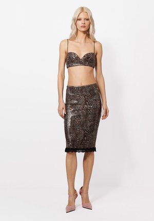 Blondynka stoi, nosząc błyszczący bralet w panterkę i dopasowaną spódnicę do kolan z czarną koronkową lamówką, połączone z różowymi szpilkami.