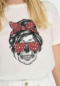 Camiseta blanca con un gráfico de una calavera que lleva unas gafas de sol rojas de gran tamaño con lunares blancos y un lazo de lunares rojos en el cabello.