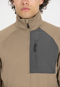 Braune, strukturierte Quarter-Zip-Jacke mit einer grauen Brusttasche. Sie verfügt über einen hohen Kragen und einen glatten Reißverschluss.