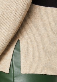 Beige gerippter Pullover mit strukturiertem Strick, der an der Säume eine schräg ausgechnitte Detail aufweist, kontrastiert mit einem grünen Lederhintergrund.