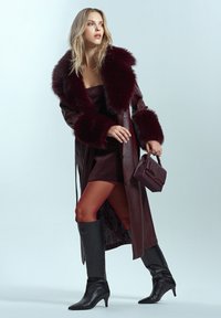 Cappotto di pelle bordeaux con colletto in pelliccia sintetica, vestito aderente, collant opachi e stivali neri al ginocchio. Borsa attaccabile con tracolla.