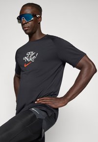 Fekete sportpóló "Fly Nike!" grafikával, narancssárga Nike logóval, fekete leggings-szel, amelyek fényvisszaverő részletekkel és testhezálló kialakítással rendelkeznek.