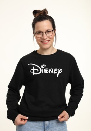 Usměvavá žena se skly a vlasem v drdolu, oblečená v černé mikině s bílým logem „Disney“ a světle modrých džínách.