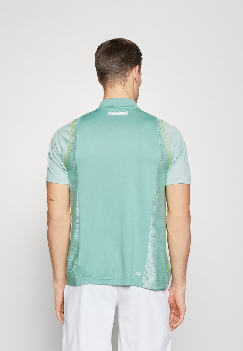 T-shirt de sport vert clair à manches courtes, avec un col, des panneaux latéraux en maille et des accents jaunes contrastés. Présente un logo au dos.