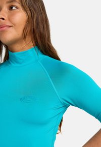 Svetlomodrý dlhý rukáv rash guard, vyrobený z elastického materiálu s krátkym golierom a diskrétnym logom na hrudi. Hladká textúra.