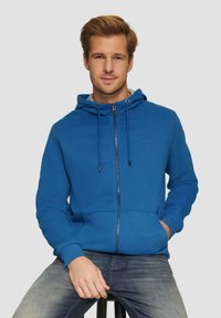 Blauwe zip-up hoodie gemaakt van zachte stof, met een trekkoordkap, voorzakken en geribde boorden. Model zit, gekleed in casual jeans.
