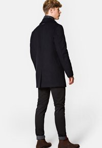 Czarny płaszcz z wełny w mieszance, o dopasowanym kroju, z kołnierzem w szpic, bocznymi wentylami i subtelnym logo na plecach. W zestawie z ciemnymi dżinsami.