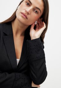 Schwarzer Blazer mit detaillierter Stickerei an den Manschetten, einem strukturierten Design und glatter Textur. Das Modell trägt ein schlichtes Oberteil darunter.