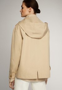 Beige jas met een capuchon, met een relaxte pasvorm, knopenaccenten en een split aan de achterkant. Gemaakt van gladde stof met een matte afwerking.