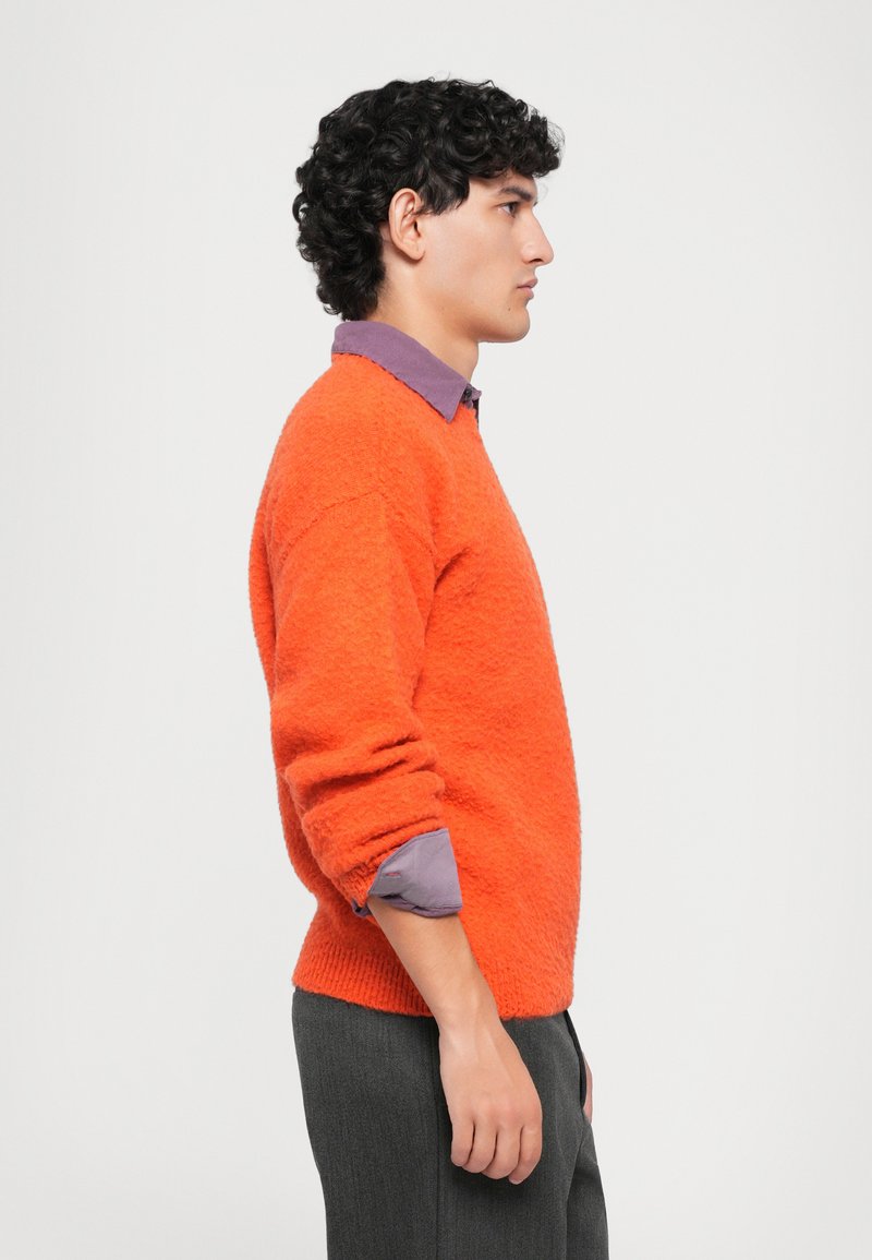 Giovane uomo con i capelli ricci che indossa un maglione arancione peloso sopra una camicia a righe viola e pantaloni grigio scuro, in piedi di lato su uno sfondo bianco.