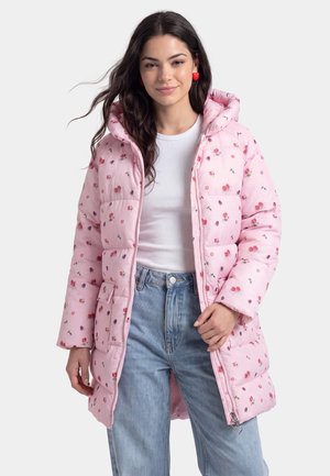 Junge Frau mit langen dunklen Haaren, die Herz-Ohrringe, eine rosa gesteppte Jacke mit Blumenmuster, ein weißes Oberteil und hellblaue Jeans vor einem einfarbigen Hintergrund trägt.