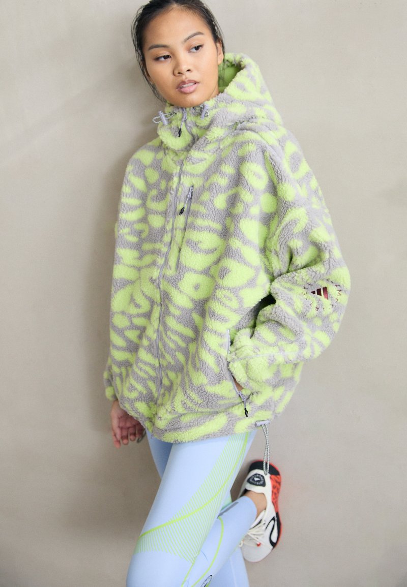 Casaco de polar em cinza com padrão abstrato em verde limão, apresentando um colarinho alto, bolsos com fecho éclair e um corte oversized.