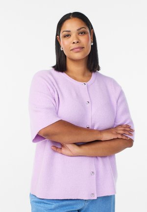 Woman with medium-length black hair wearing a lavender short-sleeve buttoned cardigan and blue jeans, arms crossed, neutral expression.