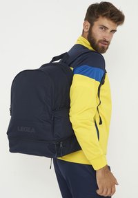 Mochila navy con una textura suave, que presenta un logotipo en la parte frontal, dos bolsillos laterales y correas ajustables. Llevada en la espalda por una persona.