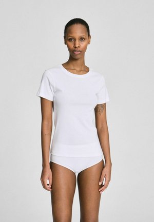 Donna con capelli corti che indossa una T-shirt bianca aderente e biancheria intima bianca, in piedi davanti a uno sfondo grigio chiaro uniforme.