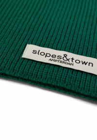 Tessuto verde a righe con forma rettangolare, caratterizzato da un'etichetta bianca che riporta la scritta "slopes&town AMSTERDAM" in testo nero.