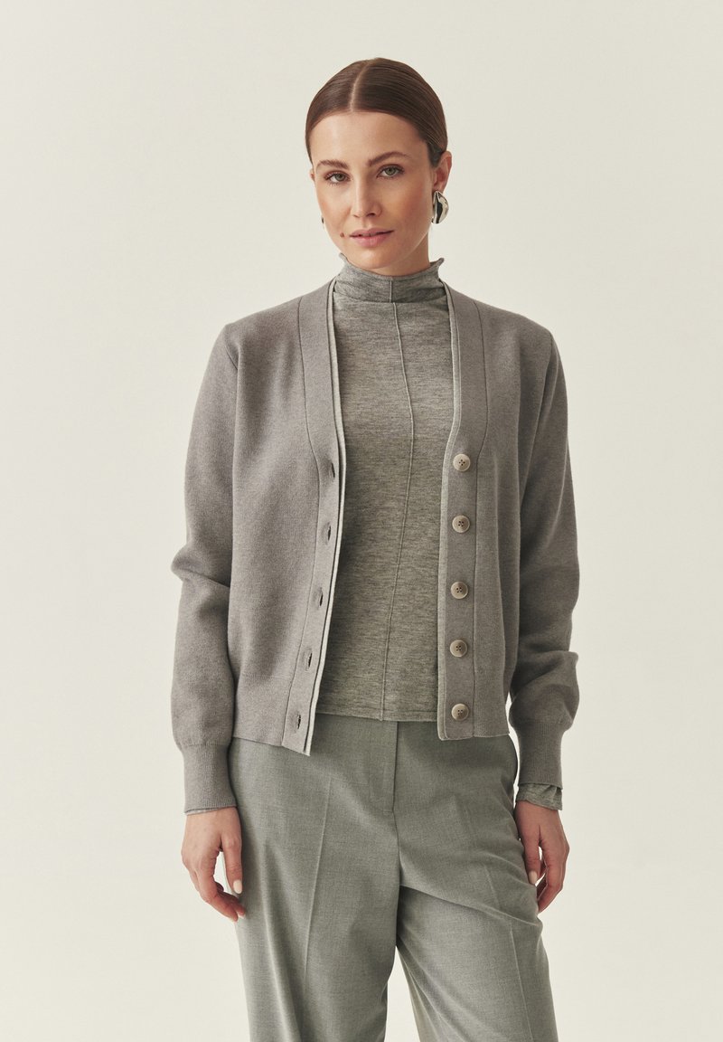 Világosszürke kardigán V-nyakkivágással és hét gombbal, szürke, álló nyakú felső fölött viselve, ugyanolyan szürke nadrággal párosítva. Puha anyagú textúra.