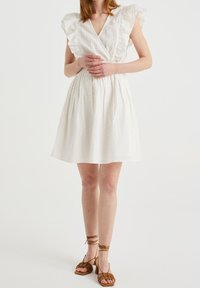 Robe blanche légère avec un décolleté en V, des manches volantes et une taille élastique. Elle présente un tissu texturé avec des motifs subtils. Sandales brunes à lanières.