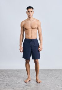 Άνδρας χωρίς πουκάμισο στέκεται ξυπόλυτος σε ένα δακτυλοειδές πατωμά φορώντας σκούρα μπλε μαγιό με κορδόνι στη μέση, κοιτάζοντας προς τα εμπρός.