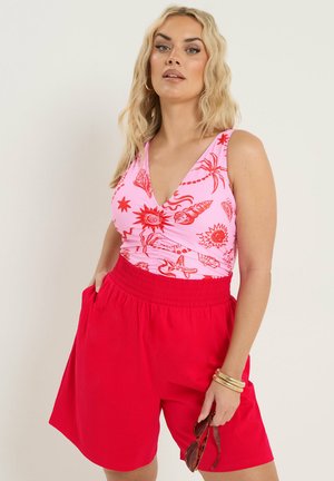 Blondynka w różowym bezrękawniku z czerwonymi nadrukami o tematyce morskiej i czerwonych szortach z wysokim stanem, trzymająca okulary przeciwsłoneczne i nosząca złote bransoletki.