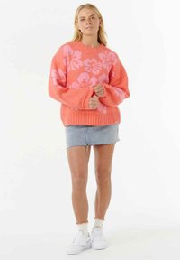 Frau in einem korallenfarbenen Pullover mit rosa Blumenmustern, einem ausgefransten Jeans-Minirock, weißen Socken mit Rüschen und weißen Turnschuhen.