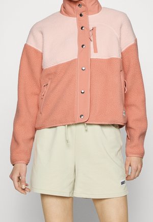 Mujer con chaqueta de forro polar rosa bicolor con botones a presión y pantalones cortos crema con cordón, sobre un fondo liso.
