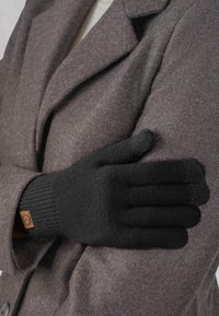 Schwarze gestrickte Handschuhe mit einem gerippten Bündchen liegen gegen einen grauen Mantel. Ein bemerkenswertes Detail ist ein kleines braunes Etikett am Handgelenk.