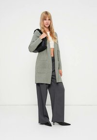 Hosszú, szürke kardigán első zsebekkel, puha anyagból készült. Széles szárú sötét nadrággal és fekete kézitáskával párosítva.