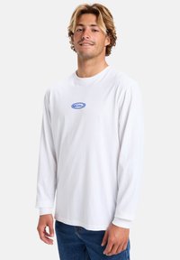 White long-sleeve t-shirt made of cotton with a round neck and blue logo centered on the chest. Paired with blue denim jeans.