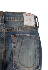 Zadná kapsa na vyblednutých modrých džínsoch True Religion s oranžovým prešívaním a bielym obdĺžnikovým štítkom značky so zobrazením Budhu hrajúceho na gitaru.