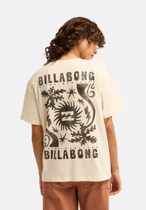Personne aux cheveux bouclés portant un t-shirt crème Billabong avec des motifs abstraits noirs représentant le soleil, des plantes et des éclairs dans le dos.