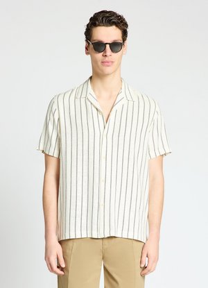 Homme portant des lunettes de soleil noires, une chemise blanche à manches courtes avec des rayures verticales noires, et un pantalon beige sur un fond clair uni.