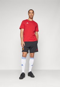 Camisola atlética vermelha com gola redonda, calções pretos com logótipo bordado, meias brancas com riscas azuis e ténis pretos.