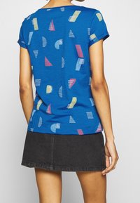 Camiseta de manga corta azul con patrones abstractos en rosa, verde y blanco. Textura suave, corte relajado, escote redondo y mangas enrolladas.