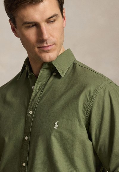 Muž pozerá dole, má na sebe zelenú košeľu na gombíky s malým bielym vyšívaným logom hráča polo na hrudi.