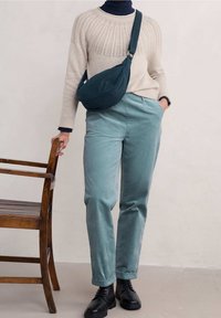 Osoba nosząca kremowy sweter, turkusowe spodnie, czarne botki i czarną torebkę na ramię, stojąca obok i trzymająca drewniane krzesło.