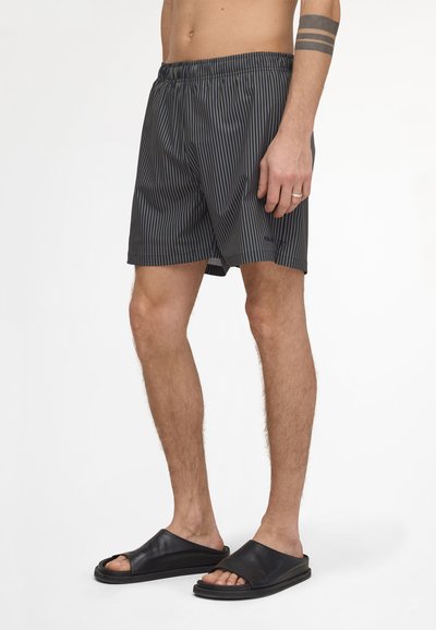 Homme portant un short rayé noir et gris avec taille élastique, sandales slides noires, montrant un tatouage et une bague à la main gauche.