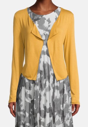Frau trägt einen senfgelben, vorne offenen Cardigan über einem grau-weißen, abstrakt gemusterten, plissierten Kleid, gezeigt von Hals bis Hüfte.