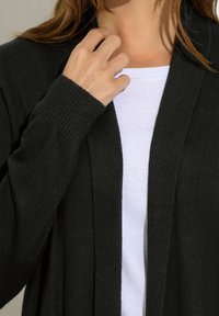 Czarny, prążkowany kardigan z otwartym przodem, z długimi rękawami i miękką fakturą, noszony na białej koszuli.