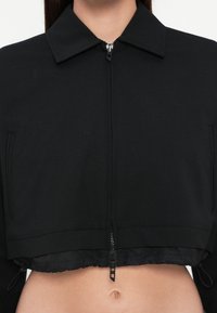 Zwarte cropped jack met een kraaghalslijn, vooraan een ritssluiting en een elastische zoom. Voorzien van zijzakken en een gladde, gestructureerde stof.