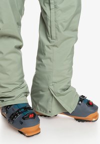 Grijze skibotten met oranje zolen, voorzien van zwarte en rode hardware, gecombineerd met lichtgroene, losse sneeuwbroeken met een verstelbare zoom.