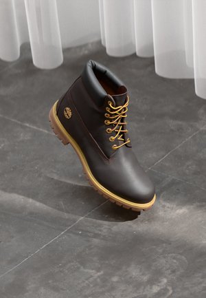 Dark brown leather ankle boot with yellow laces, padded black collar, thick beige sole, and a gold tree logo on the side, set on a gray floor.