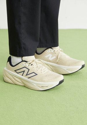 Personne portant des baskets New Balance Fresh Foam beige avec des bordures noires et des chaussettes blanches sur un sol vert clair, vêtue de pantalons noirs.