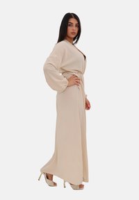 Beige lange jurk met een wikkelontwerp, ballonmouwen en een ceintuur in de taille. Gecombineerd met lichtgekleurde, met patronen versierde hoge sandalen.