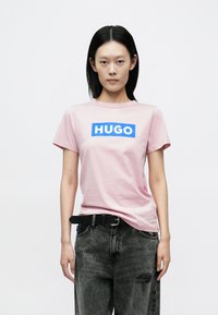 Lyserød bomuld T-shirt med en blå rektangulær "HUGO"-logo på tværs af brystet. Kombineret med mørke, slidte jeans og et sort bælte.