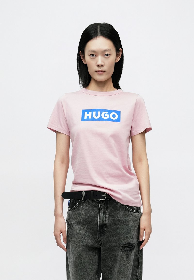 Lyserød bomuld T-shirt med en blå rektangulær "HUGO"-logo på tværs af brystet. Kombineret med mørke, slidte jeans og et sort bælte.
