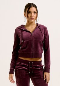 Felpa corta bordeaux con zip in un morbido tessuto effetto velluto, dotata di tasche frontali e cordini; indossata con pantaloni coordinati per un look abbinato.